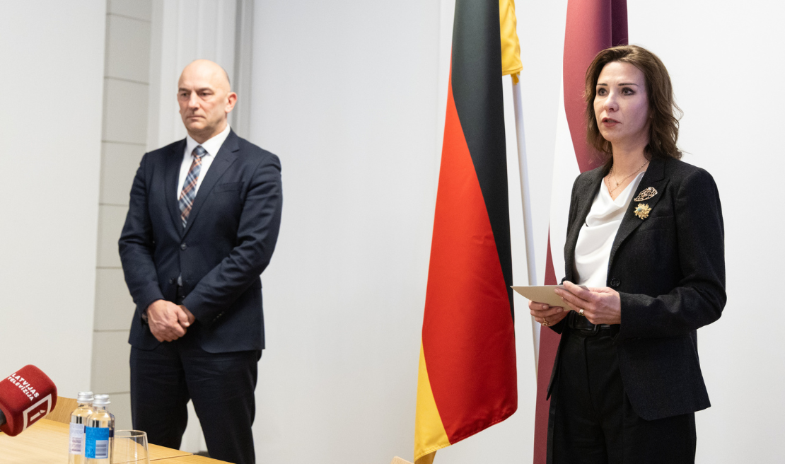 Attēlā no kreisās stāv DND vadītājs un VAK vadītāja, kas saka uzrunu pirms sadarbības memoranda parakstīšanas. Fonā ir Vācijas un Latvijas karogi.
