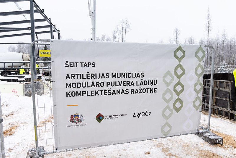 Attēla priekšpusē ir plakāts par to, ka šajā vietā taps lādiņu rūpnīca, fonā ir nedaudz redzams būvlaukums