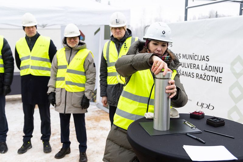 Attēla priekšpusē ir sieviete, kura skrūvē ciet ēkas pamatos iebetonējamo kapsulu. Fonā stāv vairāki cilvēki